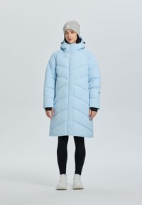 Jasnoniebieski, ocieplany płaszcz w zygzakowaty wzór, z wysokim kołnierzem i kapturem. Noszony na czarne legginsy i w zestawieniu z białymi trampkami. Szary beanie na głowie.