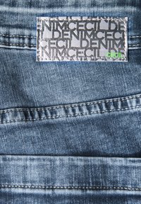 Jeans tyg i blå nyanser med en blek textur. En rektangulär etikett har ordet "Cecil" i grönt och upprepat grått text.