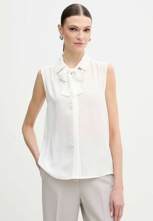 Donna che indossa una camicetta bianca senza maniche con colletto a fiocco e pantaloni beige chiaro, in piedi davanti a uno sfondo bianco uniforme.