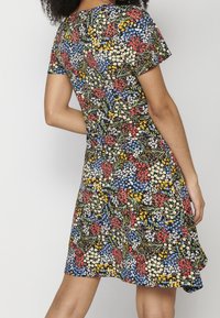 Femme portant une robe à manches courtes avec un motif floral coloré sur un fond clair, vue de dos et de la mi-cuisse vers le bas.