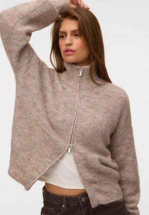 Jonge vrouw met lang haar die een beige gebreide trui met rits draagt over een wit topje en donkere broek, poserend met één arm omhoog.