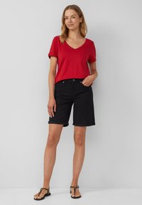 T-shirt rouge à col en V, manches courtes, associé à un short en jean noir avec des ourlets retroussés. Des sandales complètent la tenue. Fond neutre.