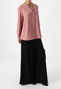 Růžová halenka s dlouhým rukávem, výstřihem do V a třemi knoflíky, v kombinaci s volnými černými kalhotami. Hladká tkanina, volný střih, minimální detaily.