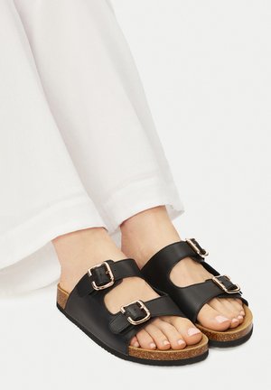Czarne skórzane sandały z dwiema regulowanymi sprzączkami, korkową wkładką i fakturowaną podeszwą, zestawione z białymi spodniami. Palce u stóp widoczne.