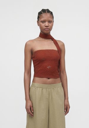 Młoda kobieta z warkoczami, ubrana w górę bez ramiączek w kolorze rdzy, ozdobioną koralikami, z dołączonym szalikiem na szyję oraz beżowe spodnie o szerokich nogawkach.