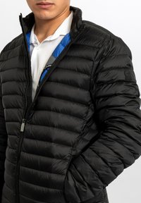 Giacca imbottita nera con collo alto, dotata di zip frontale, cuciture orizzontali e fodera interna blu a contrasto visibile al collo.