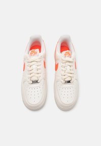 Zapatillas de cuero blancas con acentos en naranja, puntera perforada y cordones. Logo "Nike Air Force 1" en la lengüeta y en el lateral.