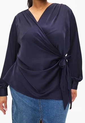Femme portant un chemisier cache-cœur à manches longues bleu marine noué à la taille, associé à une jupe en denim bleue.