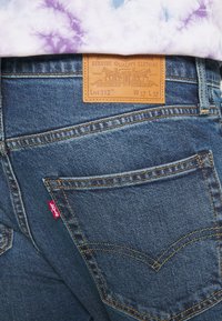 Gros plan sur la poche arrière d'un jean en denim bleu avec un patch de marque en cuir et une étiquette logo en tissu rouge, porté avec un t-shirt tie-dye violet et blanc.