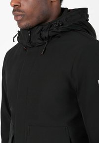 Schwarze Jacke mit hohem Kragen und verstellbarem Kapuzenzug. Besteht aus glattem Stoff, hat einen versteckten Frontreißverschluss und eine Seitentasche.