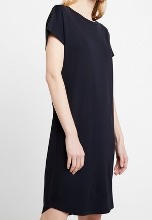 Vrouw draagt een simpele zwarte jurk met korte mouwen tot op de knie en een ronde hals op een effen witte achtergrond.