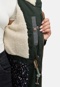 Jachetă verde cu căptușeală din blană sintetică crem, interior cu model, accesorii din bronz și șnur ajustabil. Închidere cu fermoar.