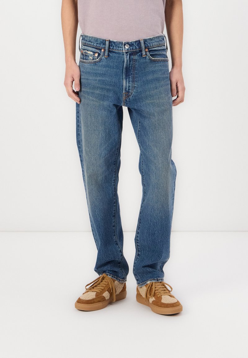 Jeans de mezclilla azul claro con corte recto, que cuentan con bolsillos frontales, un cierre de botones y un ligero desgaste en los muslos. Llevados con zapatillas color marrón claro.
