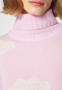 Rosa Rollkragenpullover mit geripptem Kragen und weicher Textur, mit einem weißen Wolkenmuster auf der Vorderseite. Leichtes Strickmaterial.