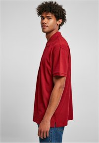 Kastanjefargad polo skjorte med klassisk krage, korte ermer og sideslitser. Laget av glatt stoff, vist på en modell i profil.