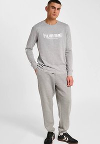 Graues Langarmshirt mit "hummel"-Logo und grauer Jogginghose. Das Outfit hat eine lockere Passform und wird mit schwarzen Turnschuhen kombiniert.