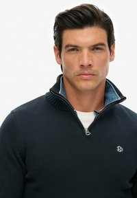 Mann mit dunklem Haar, der einen navyblauen Strickpullover über einem hellblauen Hemd mit Kragen trägt und mit neutralem Gesichtsausdruck geradeaus schaut.