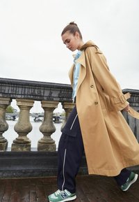 Smilgs tan krāsu mētelis ar jostu vidukļa apvidū pār gai�ši zilu denimu jakas, apvienots ar tumši ziliem kravas biksēm un zaļiem sporta apaviem, ejot gar upi.