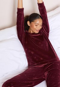Burgundské sametové domácí oblečení s dlouhými rukávy a hvězdnými vzory, s uvolněným střihem a texturovaným povrchem, na bílém pozadí.
