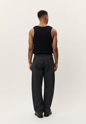 Άνδρας που στέκεται με την πλάτη στραμμένη, φοράει μαύρο αμάνικο πουκάμισο, μαύρο παντελόνι με φαρδιά πόδια και μαύρα παπούτσια σε απλό φωτεινό φόντο.