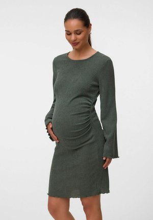 Schwangere Frau, die ein olivgrünes, langärmeliges, figurbetontes Kleid trägt, hält sanft ihren Bauch und schaut mit einem sanften Lächeln nach unten.