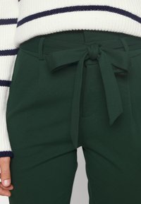 Zielone spodnie z wysokim stanem o miękkiej teksturze, z przodem związanym w kokardę i bocznymi kieszeniami, w zestawieniu z białym swetrem w niebieskie paski.