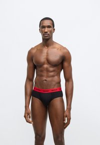 Čierne slipy s červeným pásom, na ktorom je názov značky "HUGO", vyrobené z mäkkého materiálu. Strih je priliehavý a jednoduchý.