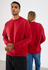 Rode sweater met ronde hals en lange mouwen, geribbelde manchetten en een effen kleur. Zichtbare stiksels en een relaxed fit. Twee modellen die van elkaar afkijken.