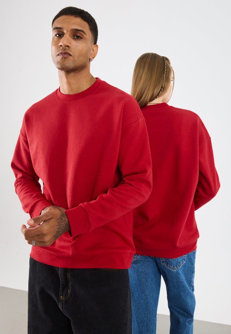 Rode sweater met ronde hals en lange mouwen, geribbelde manchetten en een effen kleur. Zichtbare stiksels en een relaxed fit. Twee modellen die van elkaar afkijken.