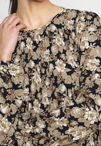 Blommig blus med svart bakgrund, prydd med krämfärgade och bruna blommor. Strukturerad tyg med veckdetaljer och långa ärmar.
