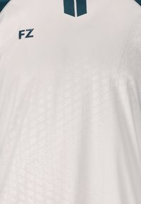 Valkoinen urheilupaita, jossa on tekstuuripintaiset yksityiskohdat, tummansiniset yksityiskohdat ja "FZ"-logo vasemmalla puolella. Pyöreä kaula-aukko ja lyhyet hihat.