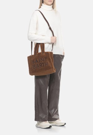Donna che indossa un dolcevita bianco e pantaloni scuri tiene una borsa a tracolla marrone pelosa con la scritta "SAINT BARTH" e una lunga tracolla sulla spalla.