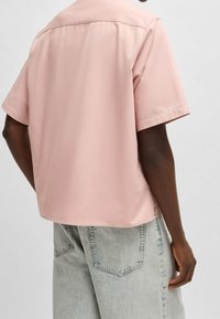 Vue de dos d'une personne portant une chemise courte rose ample et un jean en denim bleu clair avec des poches arrière visibles.