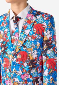 Costume coloré avec un imprimé style bande dessinée représentant des personnages sur un fond bleu, complété par une chemise blanche et une cravate à motifs.