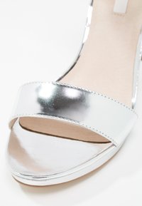 Gros plan d'une sandale argentée brillante à bout ouvert avec une large bride avant et une semelle intérieure beige clair.
