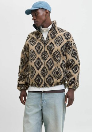 Tan fleece jacket with black diamond patterns, zip closure, high collar, and elastic cuffs. Worn over a white shirt and light denim jeans.