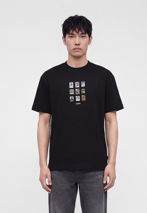 Jeune homme aux cheveux foncés porte un t-shirt noir avec une grille de neuf petites impressions graphiques colorées sur le devant, associé à un jean gris foncé.