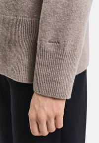 Jasnobrązowy sweter z prążkowanej wełny o gładkiej fakturze, z małym logo "GANT" na mankiecie. Dłoń opiera się o ciemną tkaninę.