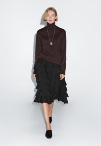 Dark brown turtleneck sweater paired with a black ruffled midi skirt. Model wears black slip-on shoes and a simple necklace. Neutral background.