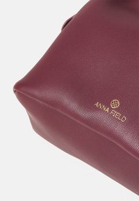 Bordo torba iz teksturirane sintetične usnjene tkanine. Ima zlato logo, angularno obliko in gladko površino z rahlo zaobljenimi robovi.