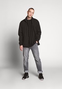 Veste légère noire sur une chemise noire à rayures, jeans gris à revers, et chaussures de sport noires avec des accents blancs.