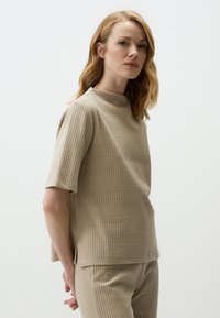 Top in tessuto beige a coste con maniche corte, colletto alto e spacchi laterali. Presenta una finitura testurizzata ed è parte di un set coordinato.