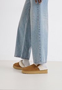 Hnědé semišové slip-on boty s bílým ovčí vlněným podšívkou, v kombinaci s světle modrými širokými džínami a bílými ponožkami, na neutrálním pozadí.