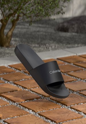 Czarny klapek Calvin Klein unoszący się nad kwadratowymi drewnianymi płytkami z małymi szarymi kamieniami na zewnątrz.