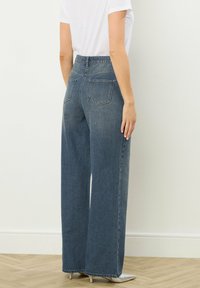 Jean en denim taille large de couleur bleu moyen. Ceinture élastique, deux poches arrière, délavage léger et détails de couture subtils. Coupe droite au niveau de l'ourlet.