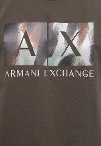 T-shirt vert olive avec un graphique texturé dans des nuances de gris, marron et vert, portant "AX" et "ARMANI EXCHANGE" en lettres grasses.