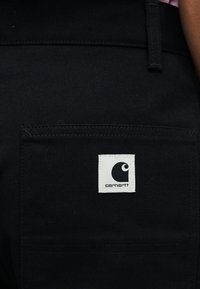 Zwarte stoffen broek met een achterzak met een wit vierkant label waarop een zwart logo en de merknaam "Carhartt" staan.