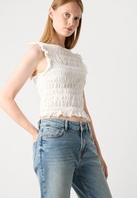 Biała bluzka ze smockiem z falbaniastymi brzegami, zestawiona z jasnoniebieskimi dżinsami. Bluzka ma teksturowany wzór i krótki krój.