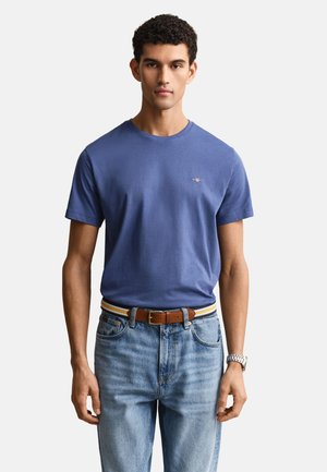 Homme debout face à l'avant portant un t-shirt bleu à manches courtes, un jean bleu clair, une ceinture marron et une montre-bracelet argentée.