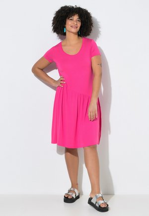 Frau mit lockigem Haar in pinkem Kurzarmkleid und silbernen Sandalen steht mit einer Hand in der Hüfte vor weißem Hintergrund.